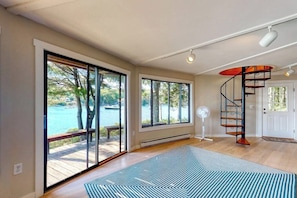 A spacious room with large windows, a spiral staircase, and a view of a body of water. - Unique Oceanfront Home With a Deck, Tons of Natural Light, & Amazing Views (Wiscasset)