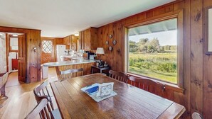 Un intérieur en bois comprenant une table à manger, des chaises, un coin cuisine et une grande fenêtre donnant sur un paysage verdoyant.