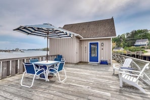 Eine Holzterrasse mit Tisch und Stühlen, eine blaue Tür und Blick auf das Wasser.