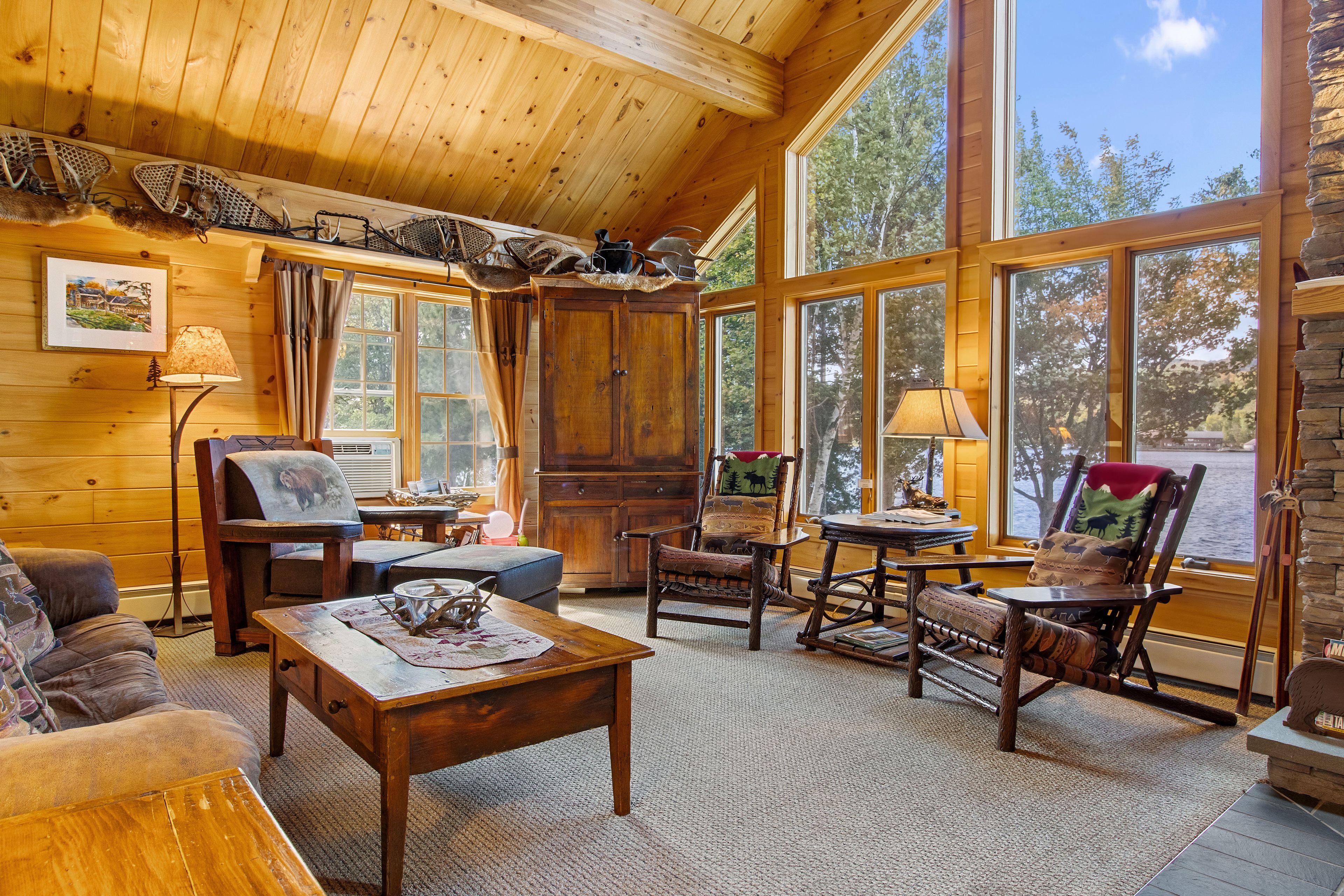 Interior acogedor de una cabaña con paredes de madera, ventanas grandes, una chimenea y muebles rústicos.