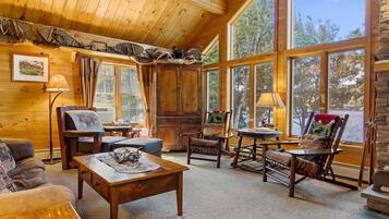 Interior acogedor de una cabaña con paredes de madera, ventanas grandes, una chimenea y muebles rústicos.