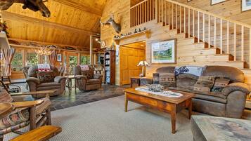 A spacious wooden interior with a high ceiling, a fireplace, a staircase, and a cozy seating area.