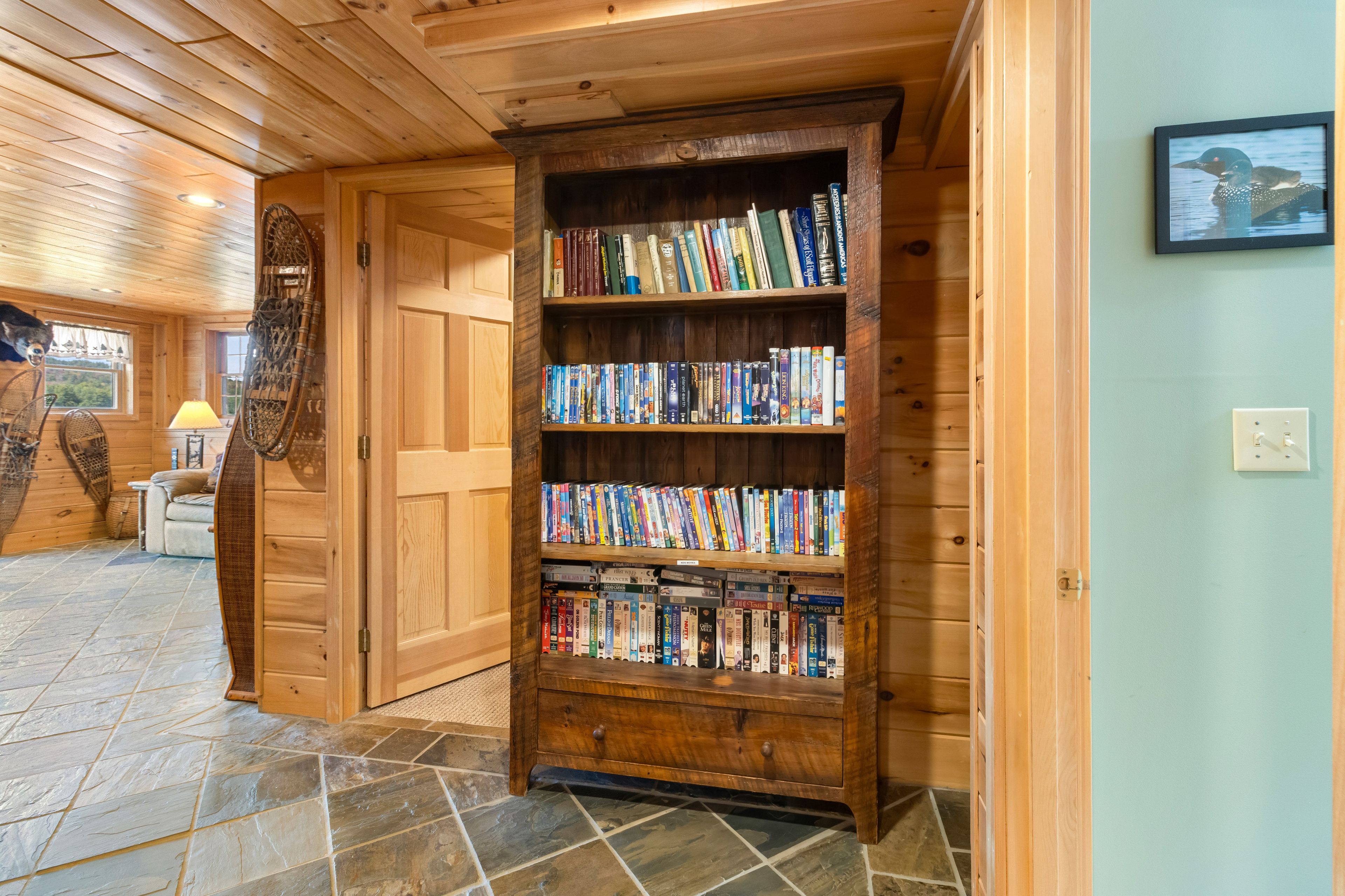 A wooden bookshelf filled with books in a room with wooden walls and a stone-tiled floor.