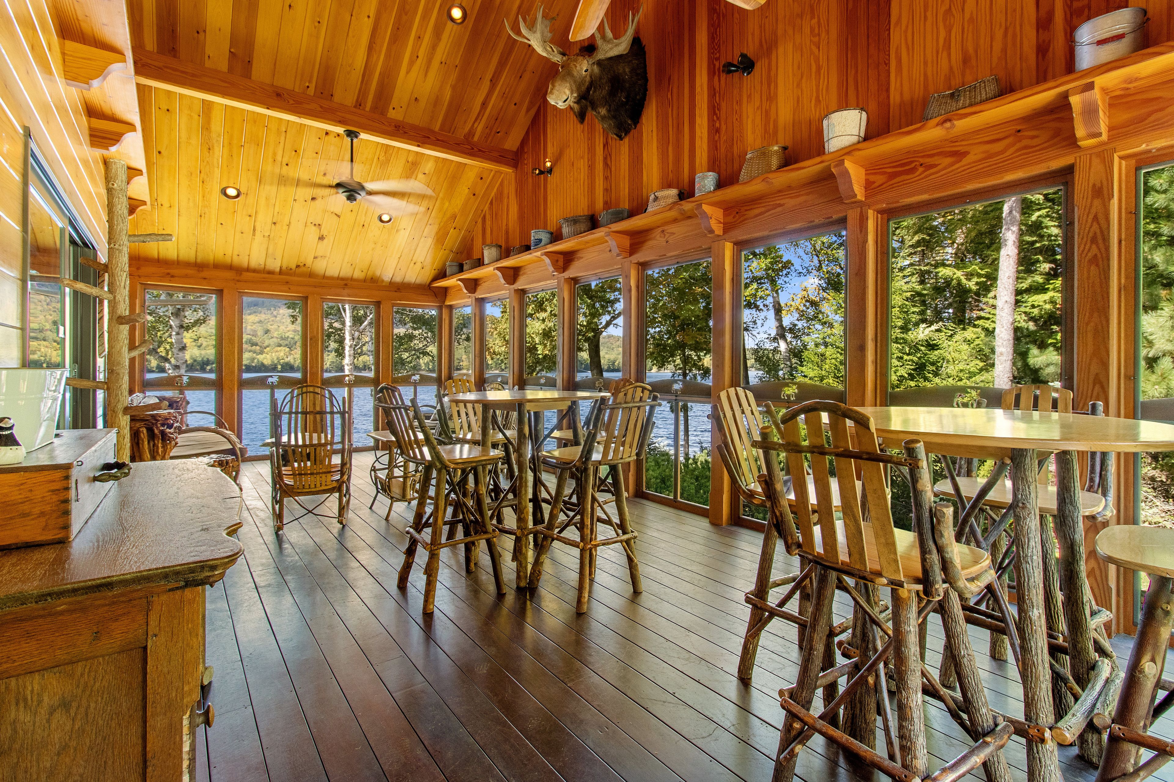 A wooden deck with rustic chairs and tables, large windows overlooking a body of water, and a ceiling fan.