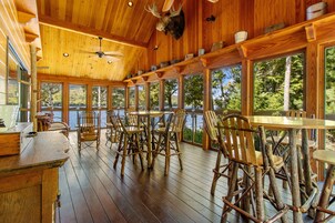 Una terraza de madera con sillas y mesas rústicas, ventanas amplias con vistas a un cuerpo de agua y un ventilador de techo.