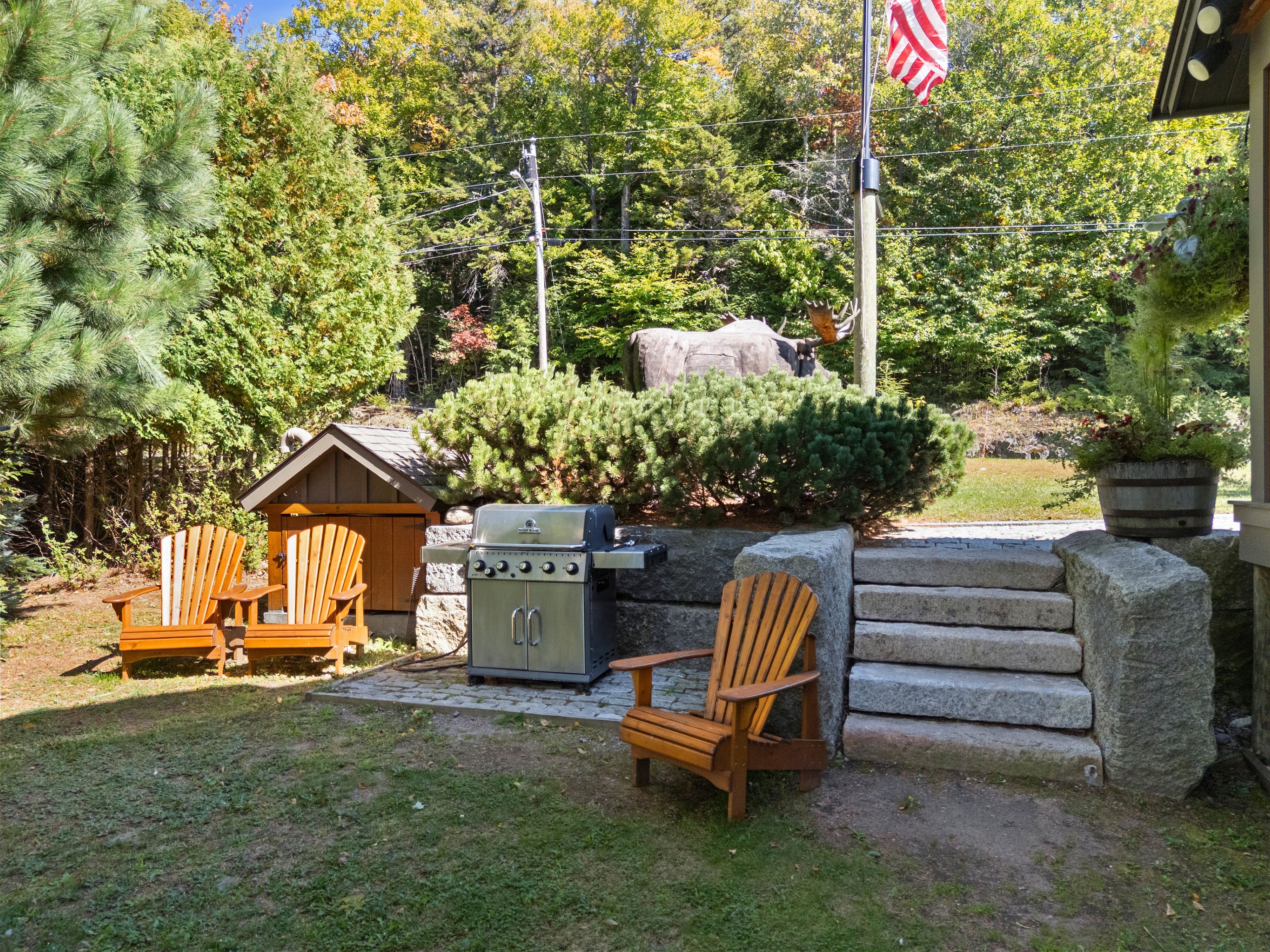 A backyard with a grill, two Adirondack chairs, a stone wall, and a flag.