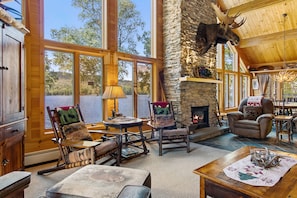 A cozy lodge interior with a stone fireplace, wooden furniture, and large windows offering a view of the outdoors. - Lakeside Lodge With Amazing Water Views, Screened Porch, Dock, & Firepit (Greenville)