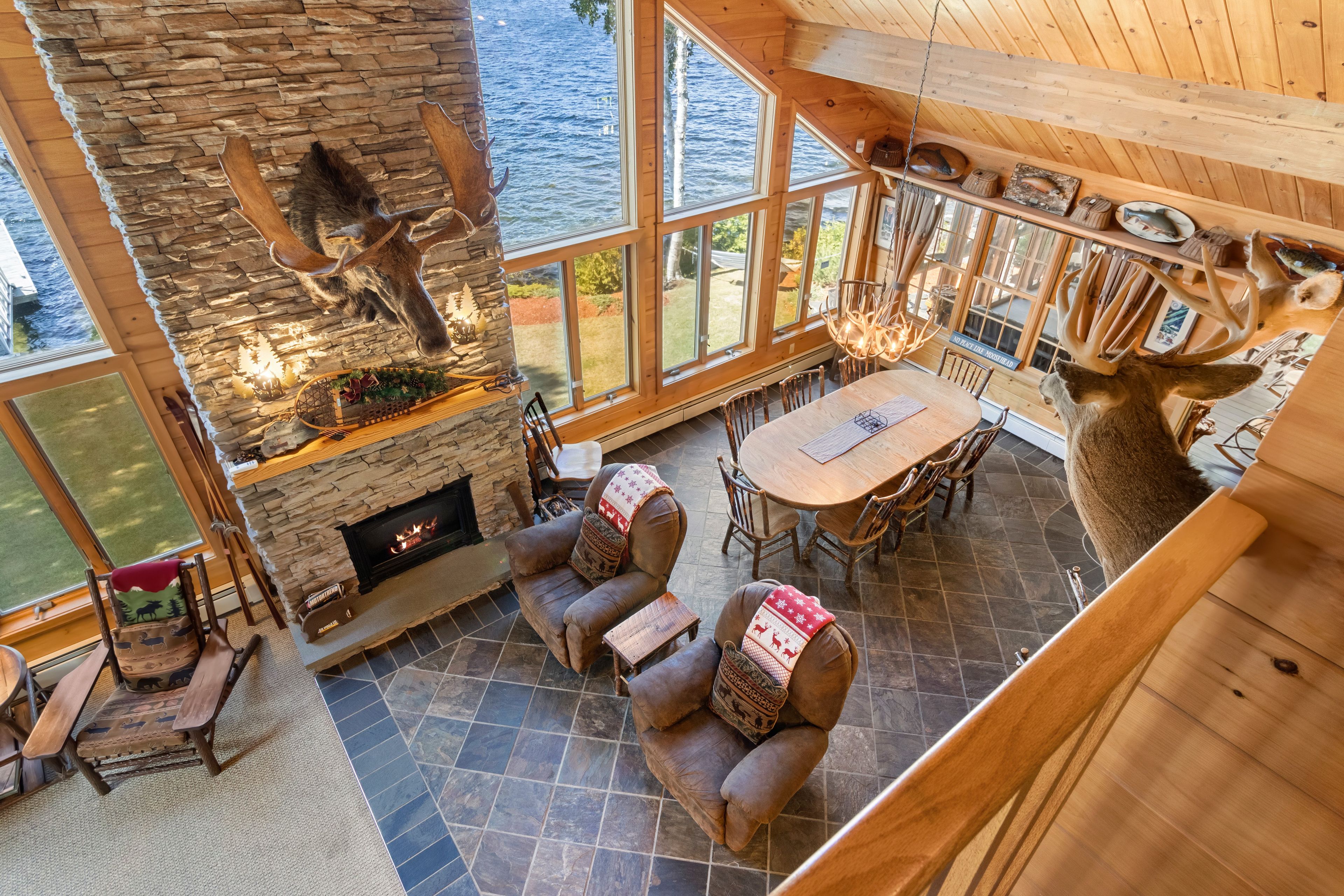 A high-ceilinged room with a stone fireplace, wooden furniture, and large windows overlooking a body of water.