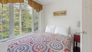 A bedroom with a large window, a patterned bedspread, a bedside table with a lamp, and a wall-mounted picture. - Ideally Located Home w/ Deck, Wifi & Washer/dryer - Walk Into Town & Acadia (Bar Harbor)