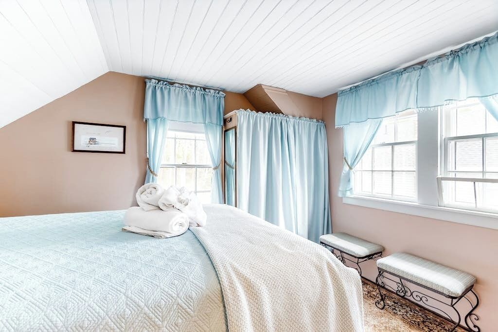 Une chambre à coucher avec un lit, un banc, un tableau encadré et une fenêtre avec des rideaux.