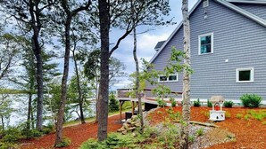 A house with a deck, surrounded by trees and a body of water. - Luxurious Waterfront Home With Deck, Firepit, hot Tub, Fireplace, & Pool Table (Boothbay)