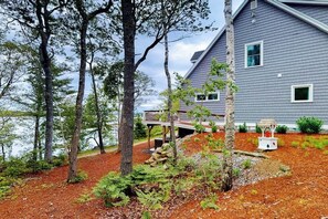 A house with a deck, surrounded by trees and a body of water. - Luxurious Waterfront Home With Deck, Firepit, hot Tub, Fireplace, & Pool Table (Boothbay)