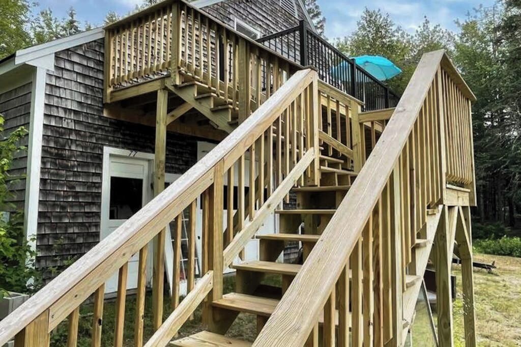 Un escalier en bois mène à une terrasse dotée d’une rambarde noire et d’un parasol bleu.