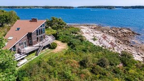 A large house with a deck overlooking a lake, surrounded by dense greenery and rocky shoreline. - Clark Point Cottage, Ocean Access, Panoramic Views (Spruce Head)