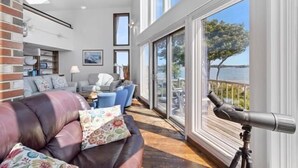 A spacious living area with a leather sofa, a dining table, and a large window overlooking a body of water. - Clark Point Cottage, Ocean Access, Panoramic Views (Spruce Head)