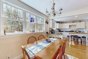A dining area with a wooden table and chairs, a chandelier, and large windows with decorative glass panes. - Coastal Bungalow With Large Yard, Beautiful Kitchen, & Great Location (Boothbay Harbor)