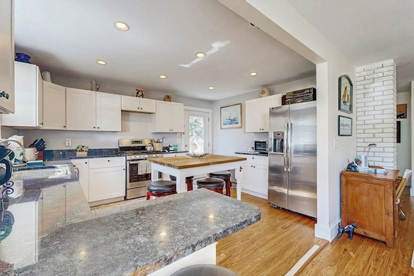 A modern kitchen with white cabinets, a granite countertop, stainless steel appliances, and a wooden dining table. - Coastal Bungalow With Large Yard, Beautiful Kitchen, & Great Location (Boothbay Harbor)