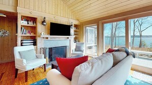 Ein gemütliches Blockhäuschen-Interieur mit Kamin, Bücherregalen, einem Fernseher und Blick ins Freie.