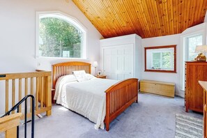 A bedroom with a wooden ceiling, a bed with a white bedspread, a window with a view of trees, a white door, and a small bedside table with a lamp. - Secluded Waterfront Home With Firepit, gas Grill, Kayaks, Dock, Balcony, & Patio (Phippsburg)