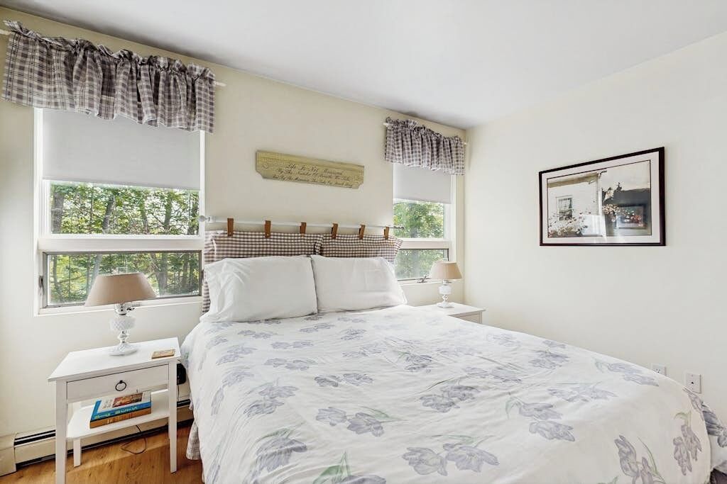 Une chambre à coucher avec un lit, deux tables de chevet, une commode, une fenêtre avec des rideaux et un tableau encadré au mur.