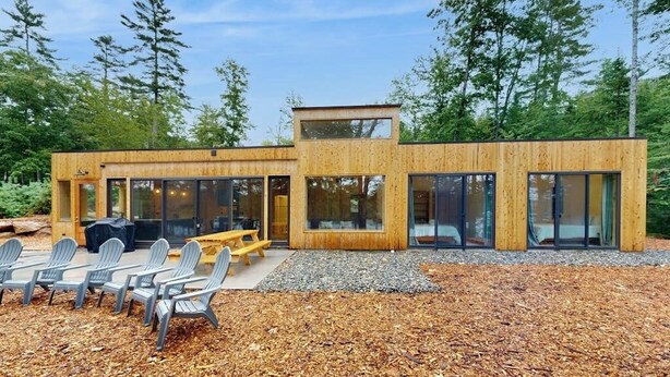 A modern wooden cabin with large glass windows, a deck with chairs, and a gravel area. - Idyllic Lakeside House With Outdoor Sauna, Patio and Firepit (Hope)