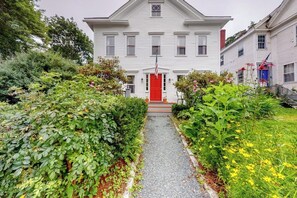 A white two-story house with a red door, surrounded by lush greenery and a gravel path leading to the entrance.
