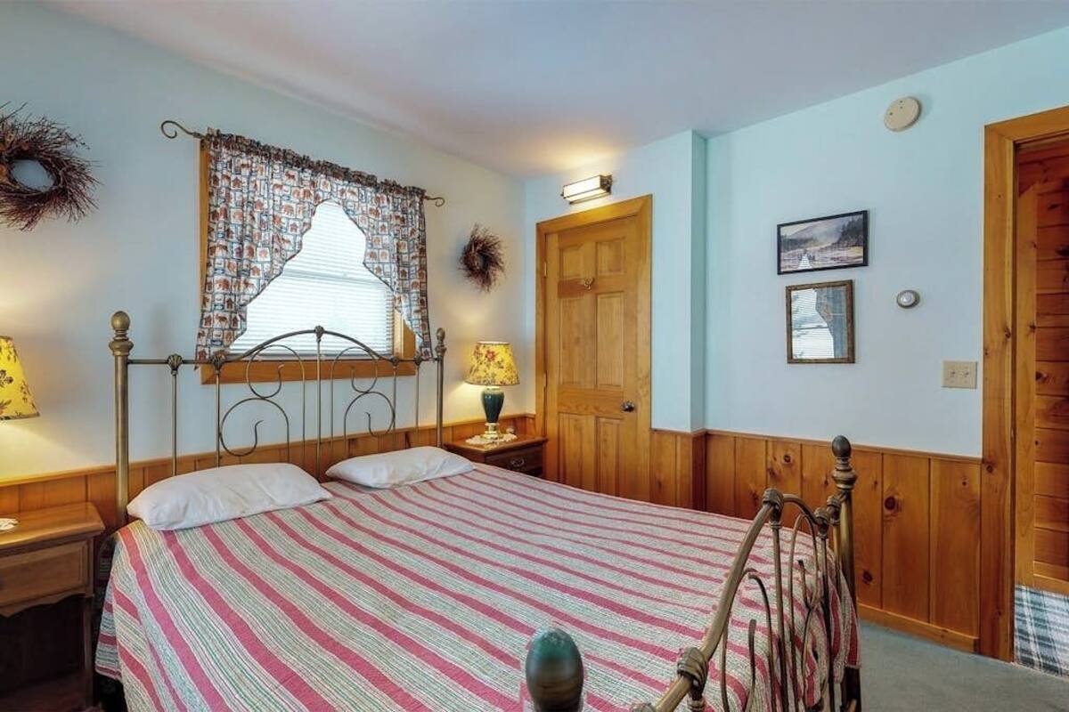 Un dormitori amb llit, panell de capçal de fusta, dues taules de nit amb làmpades, finestra amb cortines estampades, una porta i un mur amb un quadre i una corona de flors.