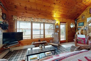 Interior de una cabaña de troncos con una televisión, una mesa de centro a cuadros, un sillón mecedor y vistas al exterior.