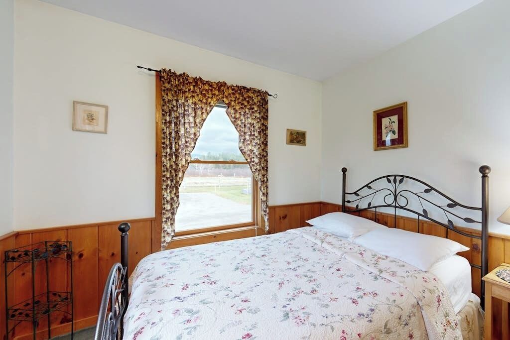 Een slaapkamer met een bed, houten lambrisering, een raam met gordijnen en een metalen hoofdbord.