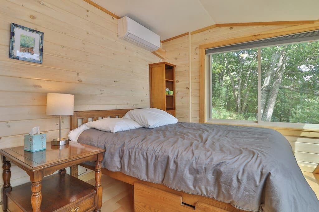 A wooden cabin bedroom with a bed, bedside table, lamp, and a window with a view of trees.