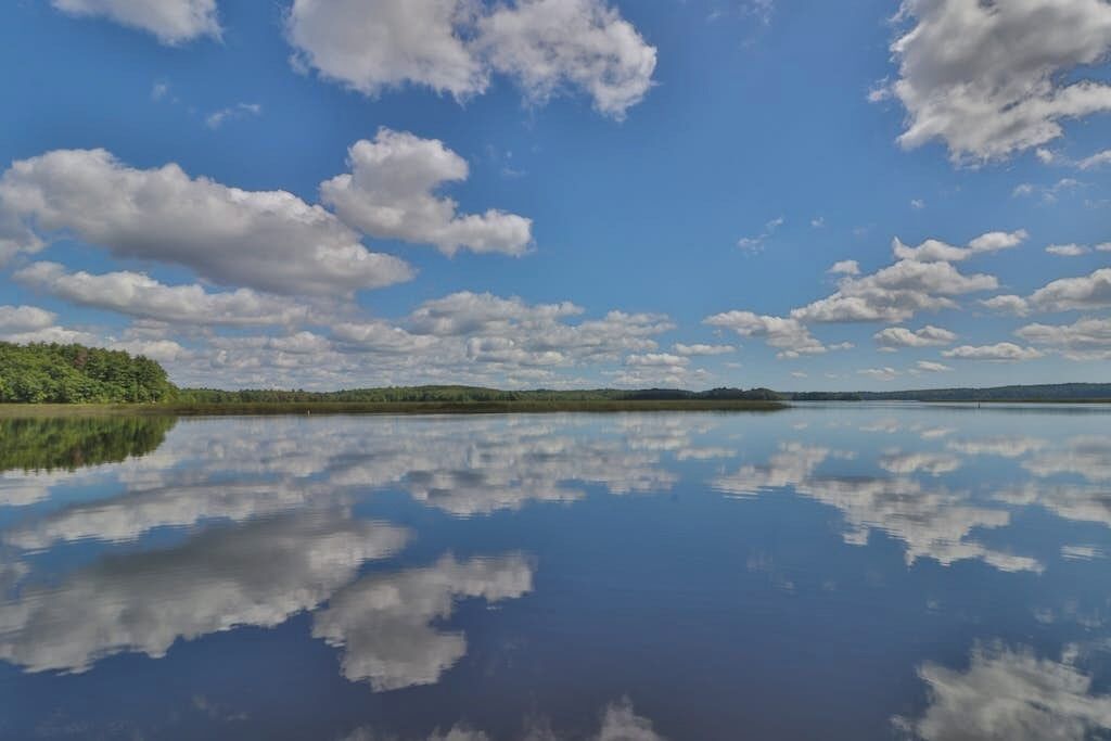 A calm lake reflecting clouds, surrounded by greenery and distant hills.