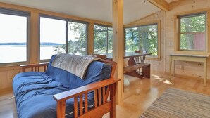 A cabin with a bed, wooden furniture, large windows, and a view of the outdoors. - Luxe Bayfront Cabin w/ Stunning Views & Shared Dock- Ideal for Hunting & Fishing (Bowdoinham)