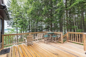 A wooden deck with outdoor furniture, surrounded by tall trees and overlooking a body of water. - Gorgeous Lakefront Home w/ Dock, Canoe, Kayaks & Lake/forest Views (Ellsworth)