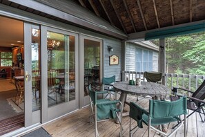 A covered outdoor deck with a wooden table and chairs, a glass door entrance, and a view of the surrounding forest. - Gorgeous Lakefront Home w/ Dock, Canoe, Kayaks & Lake/forest Views (Ellsworth)