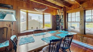 Um interior em madeira com uma mesa de jantar, cadeiras, uma estante de livros e grandes janelas.