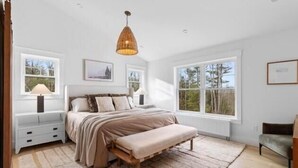 A bedroom with a large bed, a bench, a bedside table, a lamp, and a window. - Birch Tree Retreat, 6-bedroom Family Getaway Spot (South Thomaston)