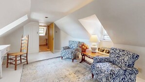 Una habitación estilo loft con techo inclinado, una silla de madera, un sillón estampado, una mesita con una lámpara y vistas por una puerta abierta.