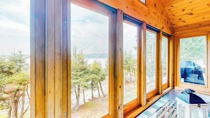 A room with large windows, wooden walls, and a view of trees and a lake. - Renovated Oceanfront Home With Stunning View & Pebbly Beach - Snowbird-friendly (Tenants Harbor)