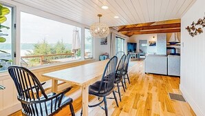 A dining area with a wooden table and chairs, a hanging light fixture, and a view through large windows. - Renovated Oceanfront Home With Stunning View & Pebbly Beach - Snowbird-friendly (Tenants Harbor)