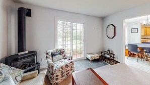 A living room with a fireplace, a floral armchair, a small ottoman, and a dining area with a table and chairs. - Idyllic Waterfront Home With Furnished Deck, Grill, Sweeping Views & Private W/D (Waldoboro)