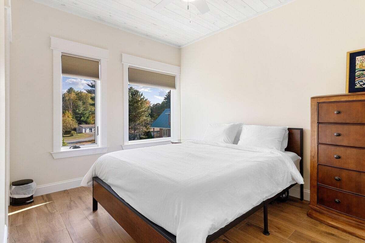 A bedroom with a bed, two windows, a dresser, and a view of trees and a house.