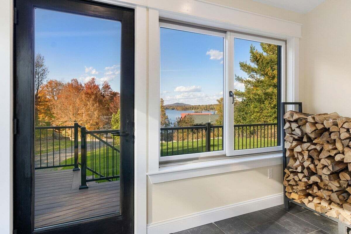 A room with a view of a lake and autumn trees, a wooden deck outside, and a stack of firewood.
