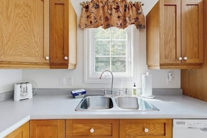 A kitchen with wooden cabinets, a double sink, a window with a floral valance, a toaster, and a dish soap bottle. - Perfect Cottage Near Downtown & Glen Mary Park w/ Full Kitchen, Free Wifi, More (Bar Harbor)