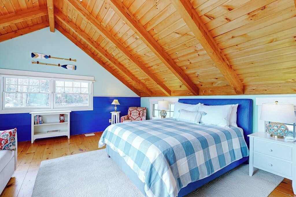 Un dormitorio con techo inclinado, una cama con colcha azul y blanca a cuadros, dos mesitas de noche con lámparas, una ventana con marcos blancos y un estante pequeño blanco con libros.