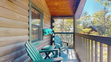 A wooden cabin with a porch, two Adirondack chairs, and a window with blinds.