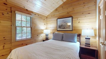 A wooden cabin bedroom with a bed, two bedside lamps, a window, and a framed picture on the wall.