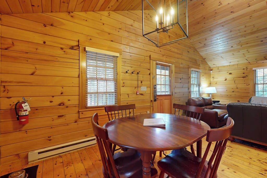 A wooden cabin interior with a dining table, chairs, a sofa, and a fireplace.