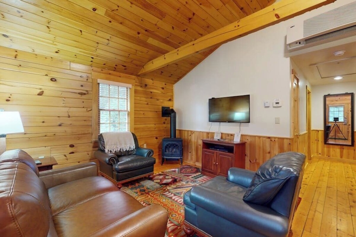 Interior de una cabaña de troncos con chimenea, sofás de cuero, televisor y techo de madera.