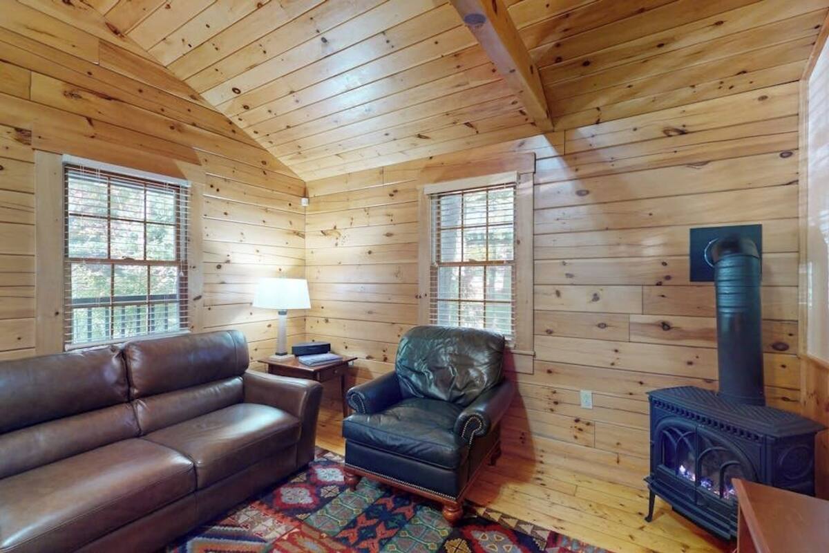 Interior de una cabaña de troncos con sofá de cuero, sillón y estufa de leña.