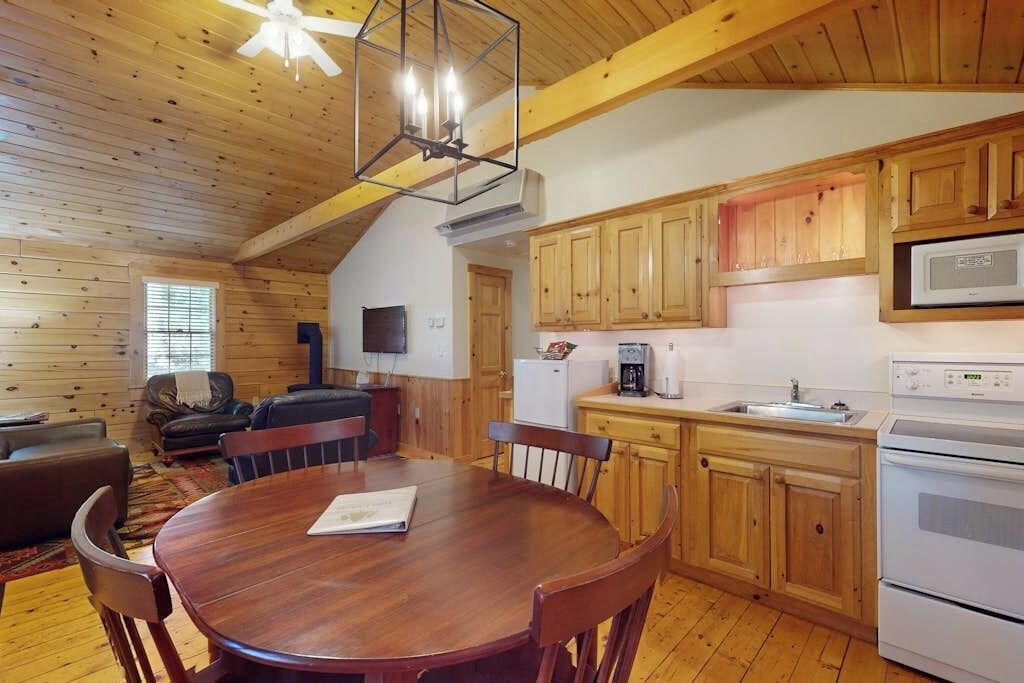 L’intérieur d’une cabane en bois comprend une table à manger, des chaises, une cuisine avec des meubles en bois, un réfrigérateur, un four à micro-ondes et un ventilateur de plafond.